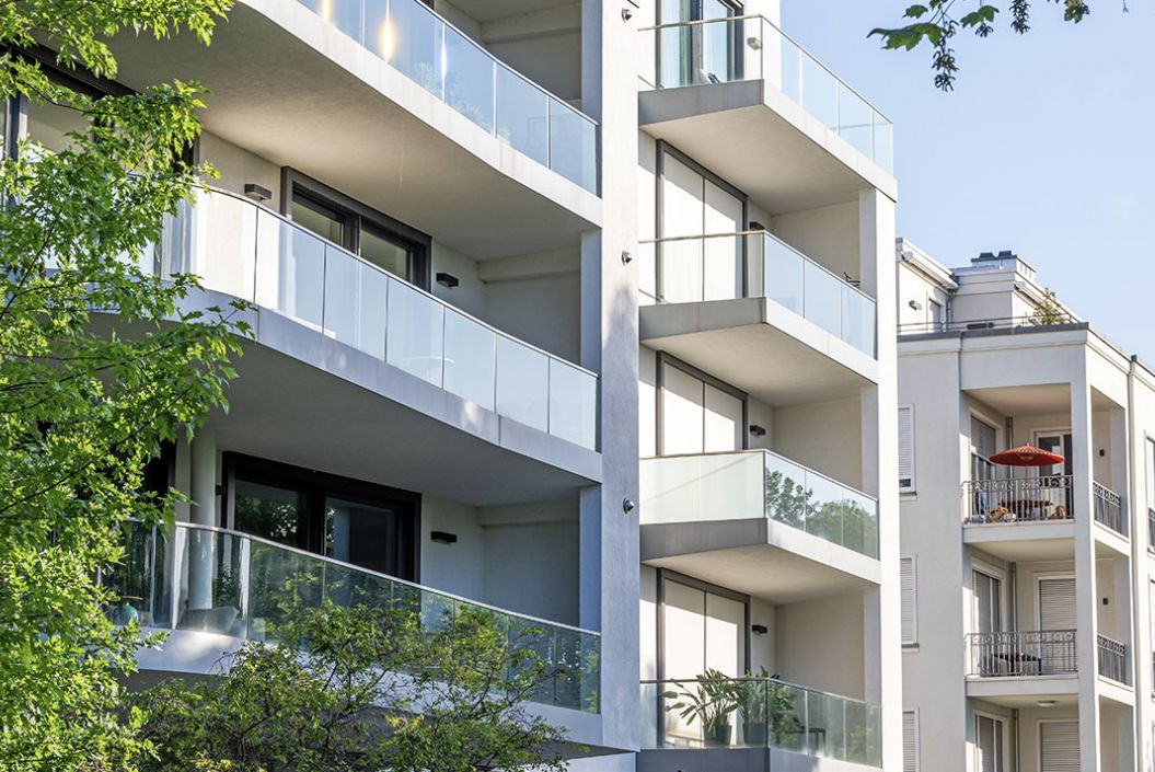 Modern apartment building with glass balconies, surrounded by trees on a sunny day.